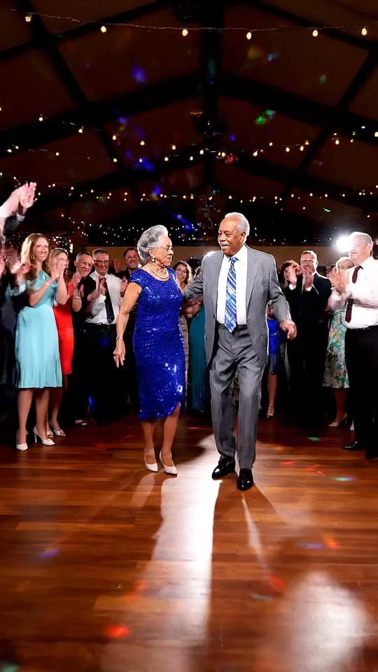An elderly couple dances rock and roll at a wedding reception while guests cheer